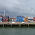 Kontenery transportowe ustawione w terminalu portowym w Rotterdamie, z dźwigami przeładunkowymi w tle.